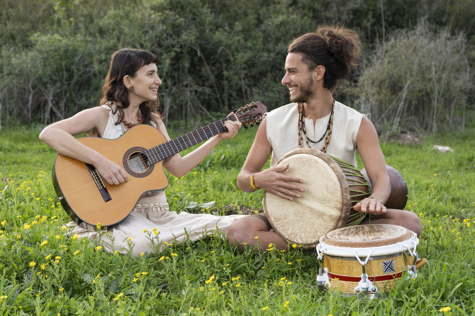 Yuli playing guitar and a bandmate playing hand drum, sitting on the grass