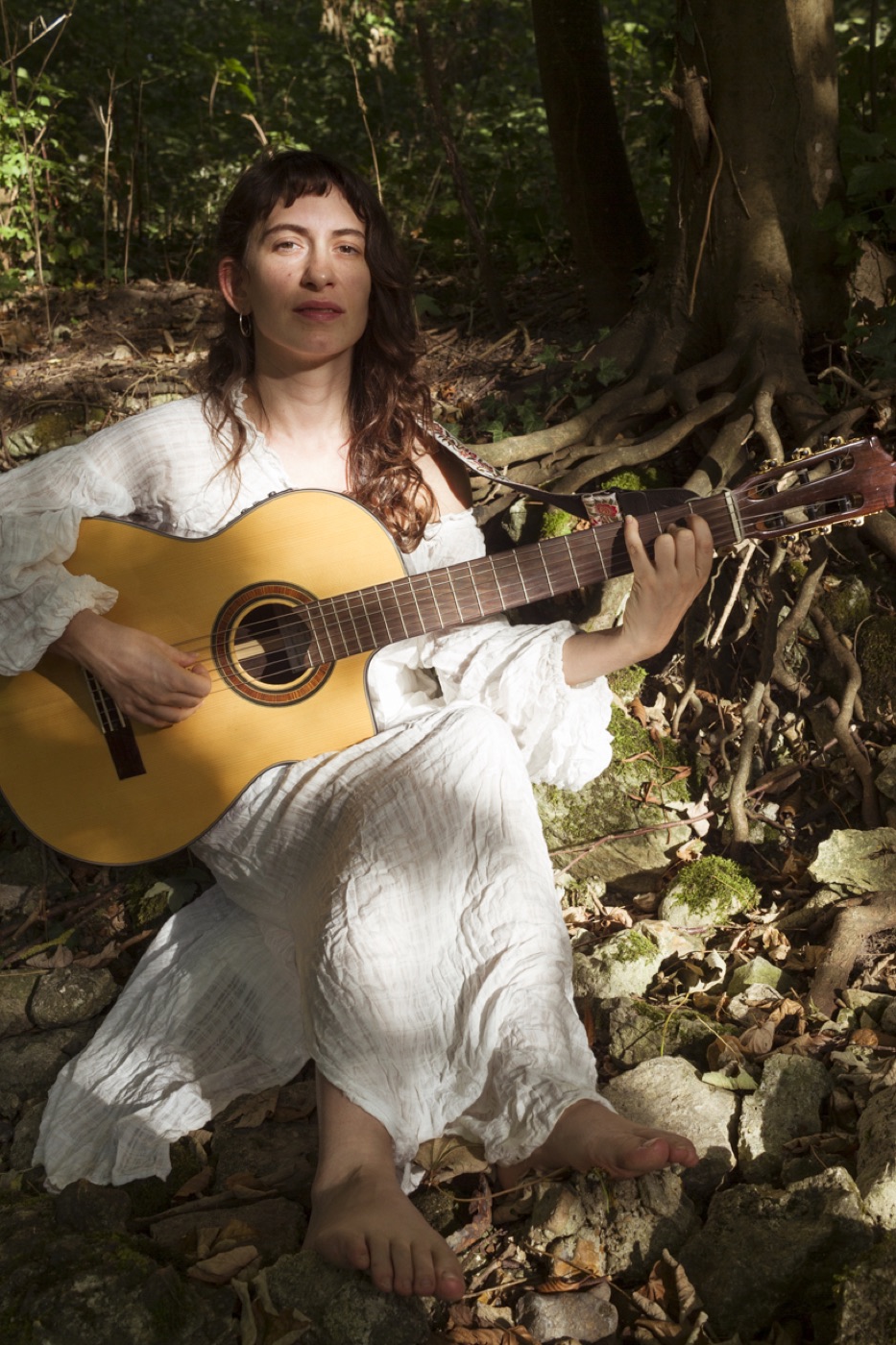 Yuli in white, sitting barefoot among tree roots in a forest, playing guitar