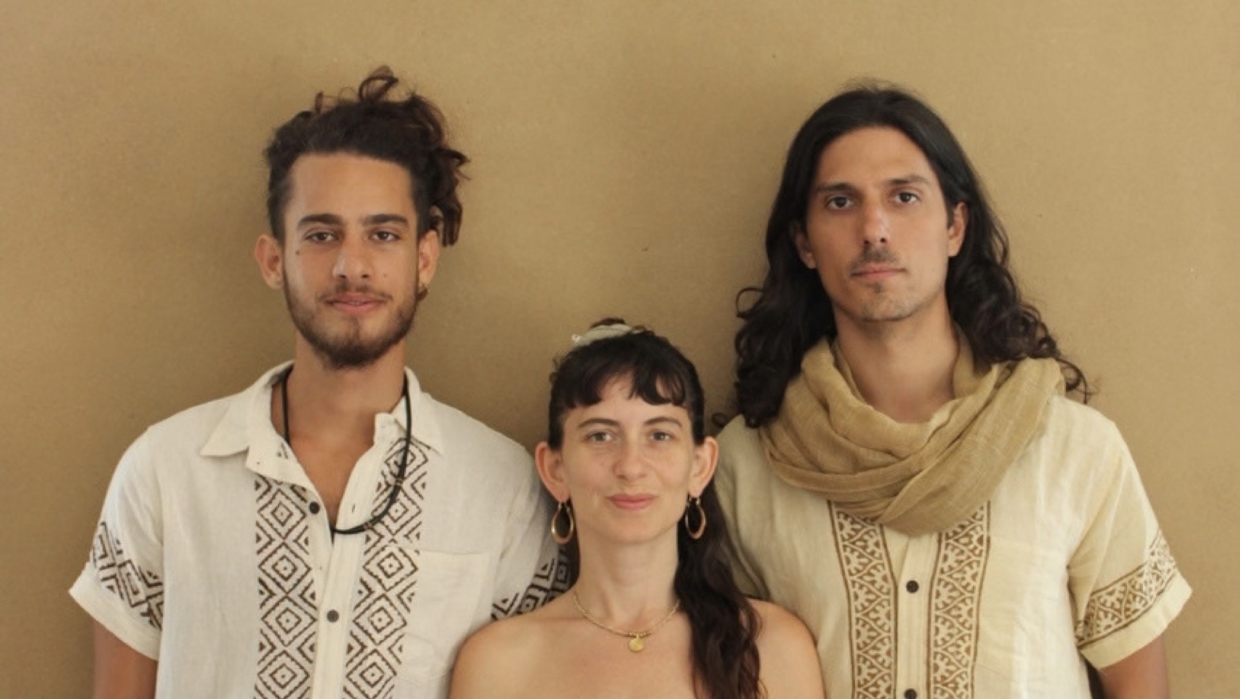 Yuli Shafriri between two bandmates in embroidered white shirts, in front of an ochre wall